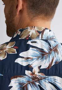 Camicetta a maniche corte con sfondo blu navy, caratterizzata da grandi motivi floreali in blu, beige e rosso. Include un'etichetta vicino al colletto.