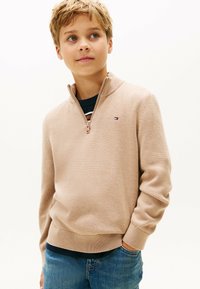 Jongen in een beige kwart-rits trui over een gestreept shirt en blauwe jeans, met een hand in de zak, die iets naar de zijkant kijkt.