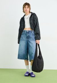 Svart mønstrert jakke, hvit kort t-skjorte, blå denimculotter, lilla skyvesandaler og en svart quiltet skulderveske.