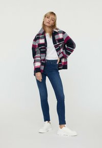 Chemise en plaid bleu marine, rose et blanc, portée sur un T-shirt blanc, associée à un jean skinny bleu et des baskets blanches. Look décontracté, coupe confortable.