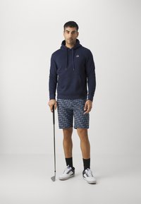 Marineblaues Hoodie mit Fronttasche, kombiniert mit gemusterten Shorts, die das Calvin Klein-Logo zeigen. Weiße Schuhe mit lila Akzenten, während ein Golfschläger gehalten wird.