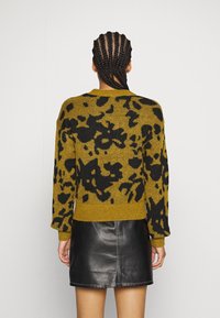 Persoon met vlechten, gekleed in een mosterdgele en zwart gepatteerde trui en een zwarte leren rok, van achteren weergegeven.