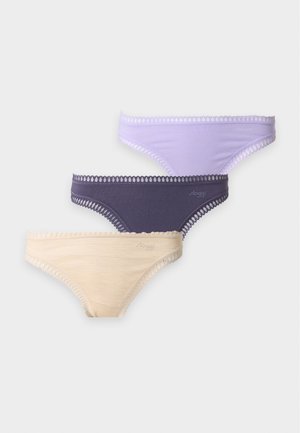 Femme aux longs cheveux foncés portant un soutien-gorge bleu sans manches et des sous-vêtements violet clair, debout pieds nus sur un fond clair uni.