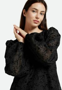 Top negro transparente con mangas abullonadas, bordado floral y puños elásticos, creando un aspecto texturizado con detalles delicados.