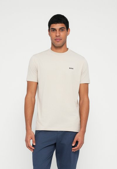 T-shirt beige à col rond en coton, avec des manches courtes et un petit logo noir "BOSS" sur la poitrine gauche. Porté avec un pantalon bleu.