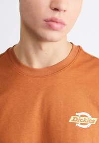 Camiseta de algodón marrón de cuello redondo con una textura suave, que presenta un pequeño logo impreso en amarillo claro y blanco en la parte izquierda del pecho.