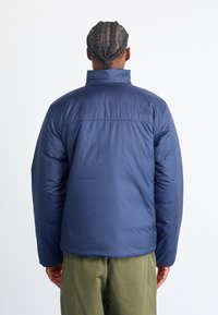Navy puffer jacket with high collar, smooth texture, and stitched detailing. Logo on upper back. Worn with olive green pants.
