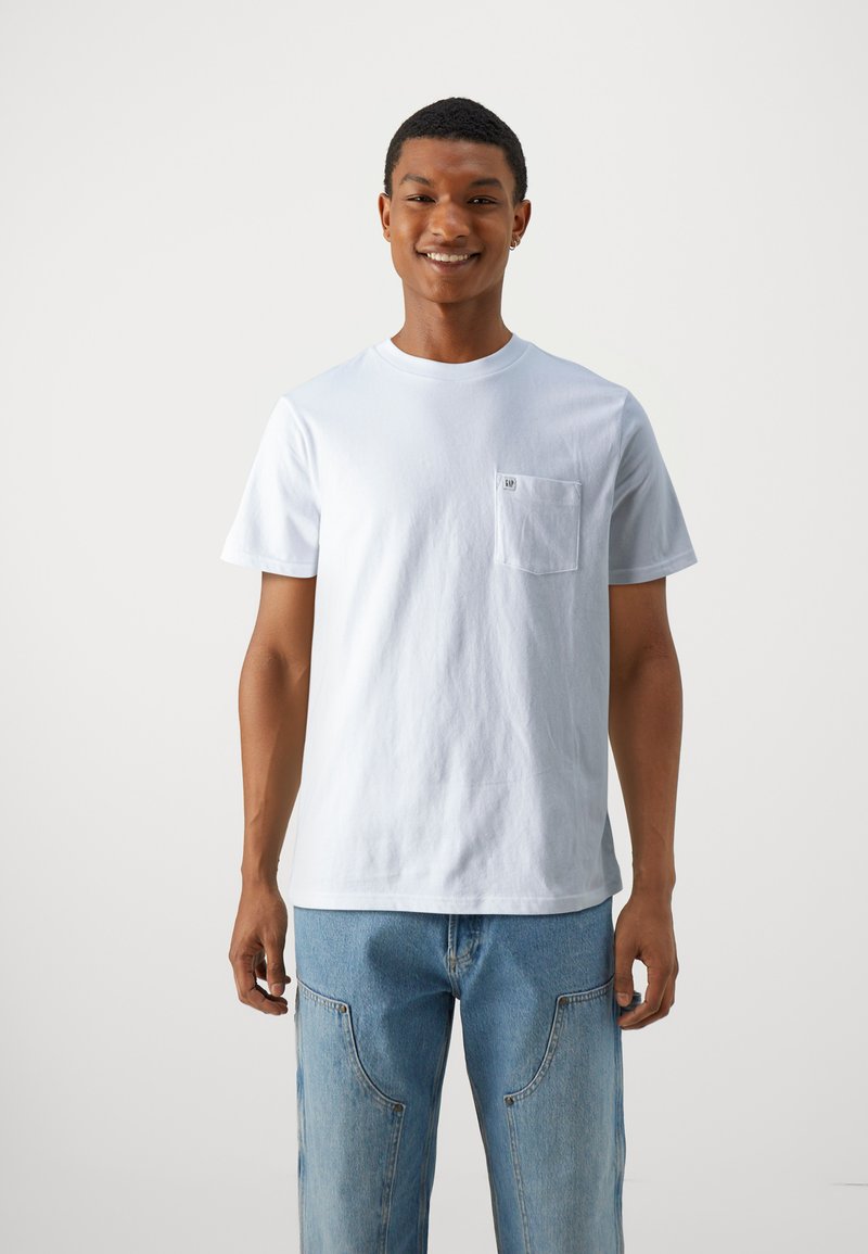 T-shirt blanc en coton avec un col rond et des manches courtes. Il comporte une poche sur le côté gauche de la poitrine et une coupe décontractée. Porté avec un jean en denim bleu clair.