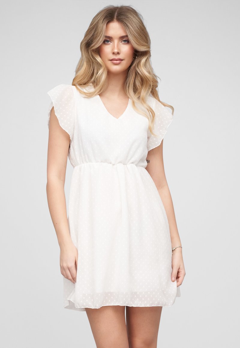 Jeune femme aux longs cheveux blonds portant une robe blanche à manches courtes avec des manches transparentes à pois sur un fond clair uni.