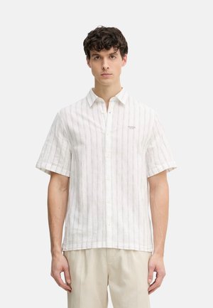 Młody mężczyzna w białej koszuli z krótkim rękawem z cienkimi pionowymi paskami i beżowych spodniach, stojący na jednolitym tle.