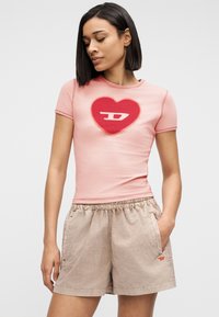 Γυναίκα με κοντά σκούρα μαλλιά φορώντας στενό ροζ μπλουζάκι με κόκκινη καρδιά και μπεζ σορτς με ελαστική μέση, στέκεται με το ένα χέρι στην τσέπη.