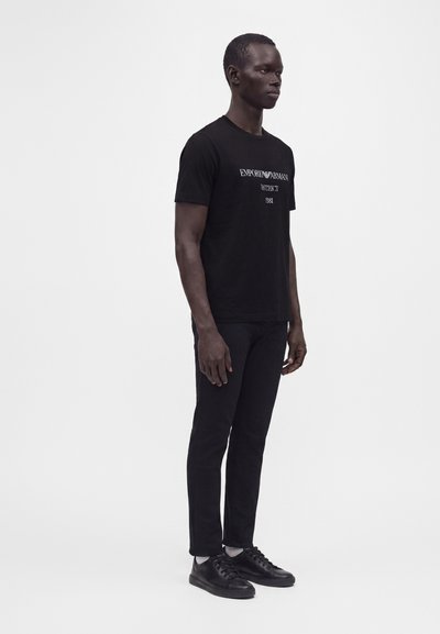 Άνδρας που φοράει μαύρο t-shirt Emporio Armani, μαύρο παντελόνι και μαύρα αθλητικά παπούτσια, στέκεται προφίλ μπροστά σε απλό λευκό φόντο.