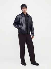 Jeune homme portant une veste en cuir noire, une chemise noire, un pantalon ample sombre et des chaussures noires, debout devant un fond clair uni.