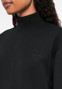 Frau trägt einen schwarzen Rollkragenpullover mit "GUESS NINETEEN EIGHTY ONE" auf der Brust gestickt.
