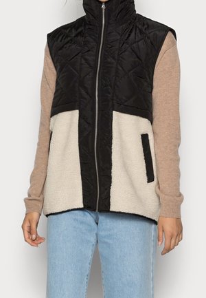 Person iført en ærmeløs sort quiltet og cremefarvet sherpa-vest med lynlås over en beige langærmet top og lyseblå jeans.