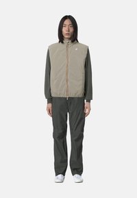 Gilet imbottito beige chiaro con colletto alto, chiusura con zip e striscia multicolore; abbinato a pantaloni verde scuro e sneakers bianche.