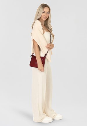 Beige gerippter Cardigan, cremefarbener Jumpsuit, roter Leder-Crossbody-Bag, weiße Sneakers, stehend vor einem hellgrauen Hintergrund.