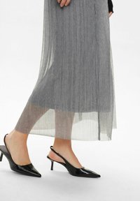 Svarta lackade slingback-klackar med spetsig tå och en liten klack, kombinerade med en grå plisserad kjol med en subtil glans.
