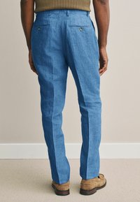 Lichtblauwe linnen broek met een rechte snit, voorzien van twee achterzakken met knopen, gedragen met een paar bruine suède schoenen.