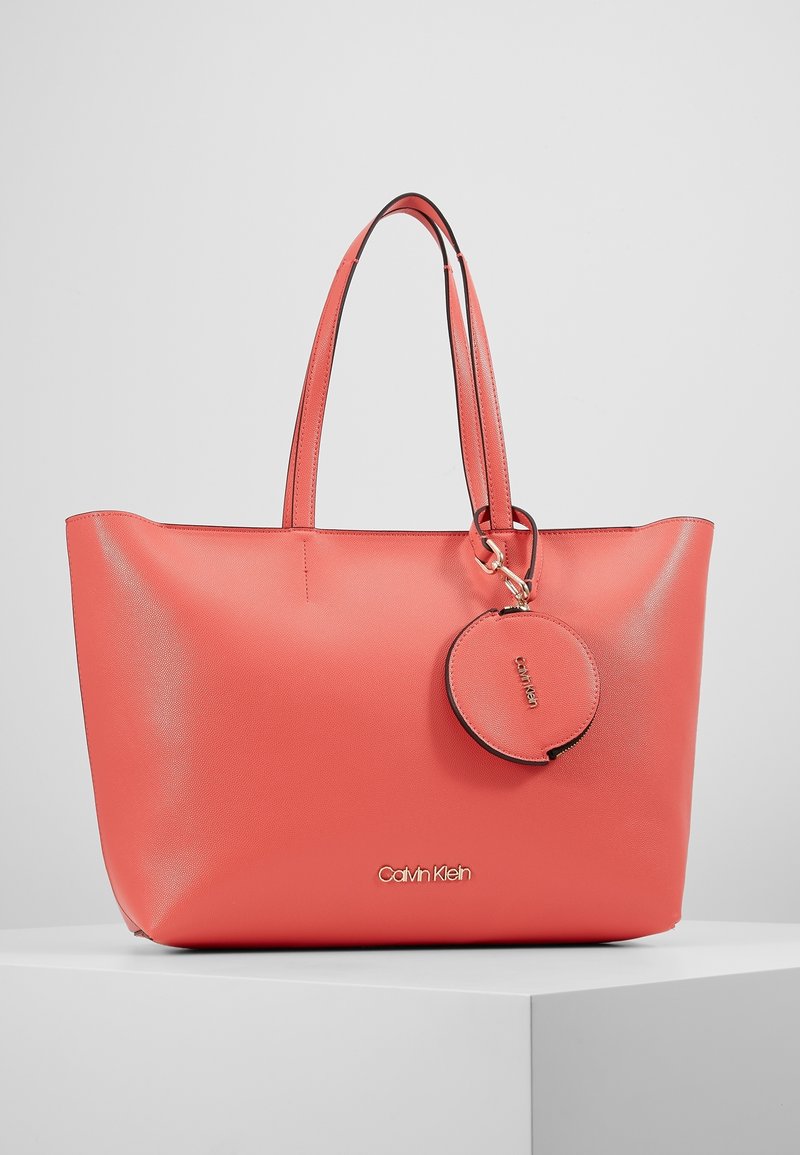 Sac cabas en cuir corail avec deux longues poignées, équipé d'une étiquette ronde pour bagages et d'un accent de marque en bas. Texture lisse, design minimaliste.