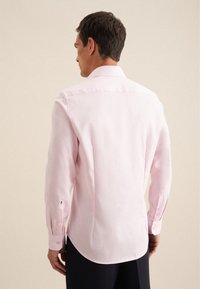 Camicia a maniche lunghe rosa chiaro con colletto classico. Presenta polsini con bottone e orlo curvo. Texture liscia con dettagli di pieghe sul retro.