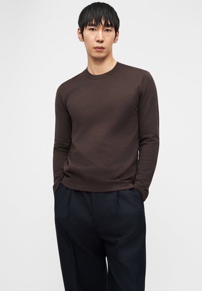 Νεαρός άνδρας με τα χέρια στις τσέπες, φορώντας σκούρο καφέ μακρυμάνικο πουκάμισο και σκούρο ναυτικό πτυχωτό παντελόνι, σε λευκό φόντο.