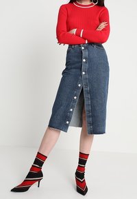 Jupe en denim avec une fermeture à boutons sur le devant, fente haute et ourlet brut. Associée à un pull côtelé rouge et des chaussettes à talons rayées.