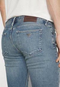 Blå denimjeans med smal passform, med en brun läderlogotyp på baksidan av midjan och en silveremblem på högra fickan.