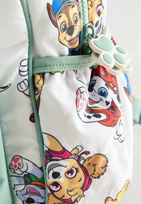 Sac à dos en tissu blanc ornée de chiens de dessin animé colorés. Agrémenté de finitions vert menthe et d'un tirette de zip en forme de patte.