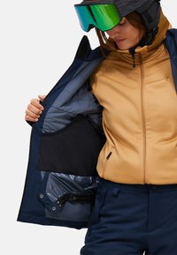 Person trägt einen Skihelm und grüne, reflektierende Goggles und zeigt die Innentasche einer navyblauen Skijacke über einem hellbraunen Reißverschluss-Hoodie.