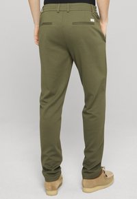 Pantalon vert olive avec une texture lisse, doté de deux poches arrière et d'une petite étiquette de marque discrète. Porté avec des chaussures à enfiler marron clair.