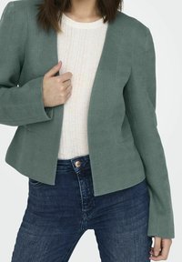 Veste verte texturée à motif chevron, à l'avant ouvert et aux manches allongées, portée par-dessus un haut côtelé de couleur claire et un jean foncé.