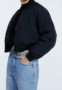 Zwarte cropped bomberjack met een ritssluiting aan de voorkant, geribbelde manchetten, gewatteerde textuur en een zakje aan de voorkant. Gecombineerd met licht denim jeans.