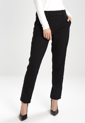 Femme portant un pantalon noir tailleur avec un détail sur le côté, un haut blanc à manches longues et des chaussures à talons hauts pointues noires, debout.
