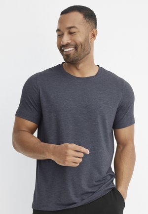 Smiling man with short hair and beard wearing a fitted dark gray t-shirt, one hand in pocket and the other slightly raised.
