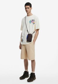 Maglietta a maniche corte di colore crema con toppe colorate, abbinata a pantaloni corti beige e stivaletti scuri. Una piccola borsa a tracolla marrone con dettagli.