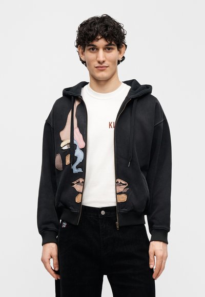 Μαύρο φούτερ με φερμουάρ και πολύχρωμο κεντητό αφηρημένο σχέδιο προσώπου, με κουκούλα με κορδόνι και ριμπ μανσέτες. Φορεμένο πάνω από λευκό t-shirt.