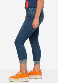 Slim-fit farkkukankaat sinisävyiset, keski korkealla vyötäröllä ja käännetyillä lahkeilla, yhdistetty kirkkaan oransseihin nauhakenkiin, joissa on teksturoitu pohja.