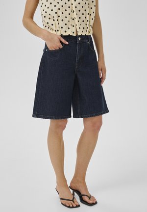 Vrouw draagt donkerblauwe denim shorts met wijde pijpen, beige mouwloze top met zwarte stippen, en zwarte sandalen met hakken, staat met één hand in de zak.