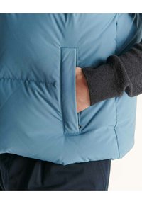 Lichtblauwe pufferjas met een gestructureerde grijze manchette. Heeft een zijzak geïntegreerd in de jas voor handige opslag.