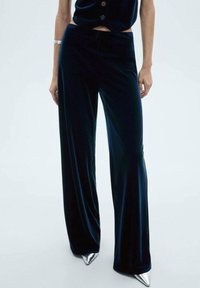 Samettileveät housut syvässä tummansinisessä värissä, joissa on sileä tekstuuri ja rento istuvuus. Yhdistetty hopeisiin kärkipohjallisiin korkokenkiin kontrastiksi.