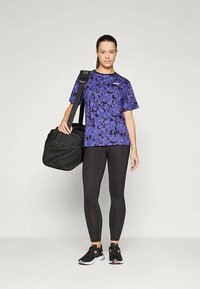 Purpura rakstains sporta T-krekls, melni legingi un melni skriešanas apavi. Tur rokās melna sporta soma ar teksturētu virsmu. Taisnstūra dizains.