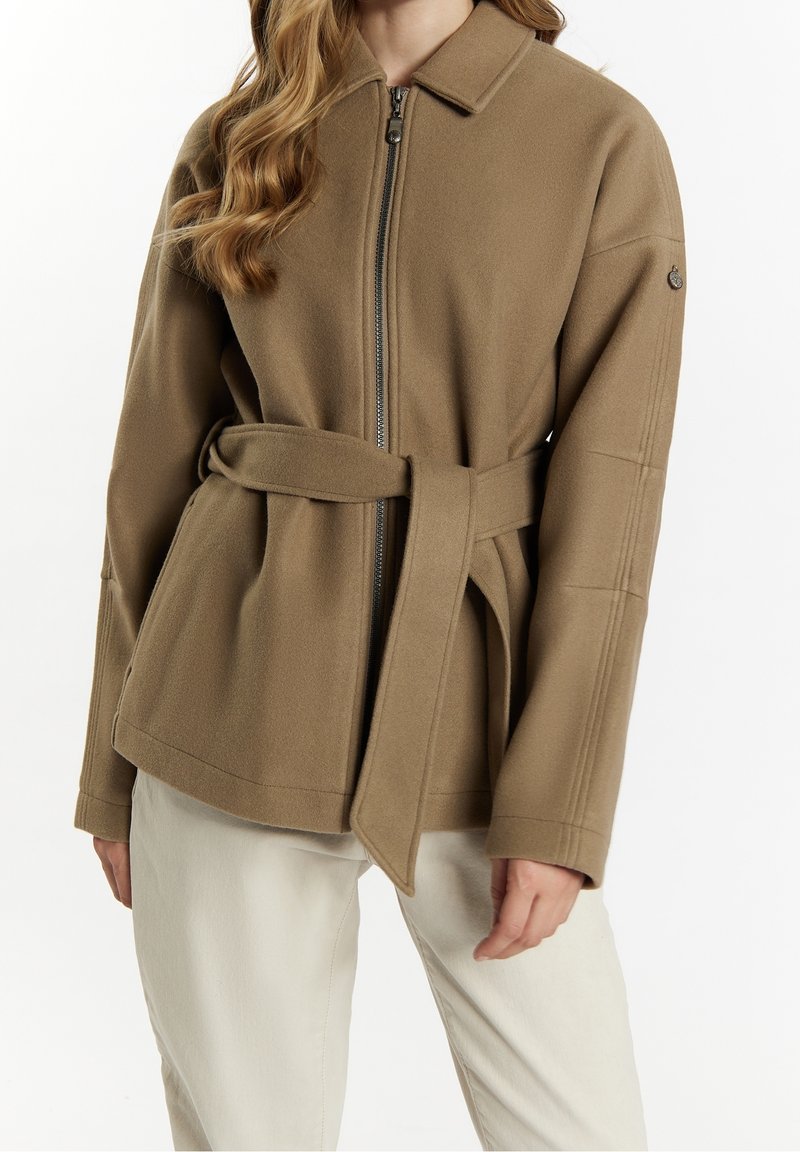 Femme portant une veste camel ceinturée avec une fermeture éclair devant et un col, associée à un pantalon beige clair.