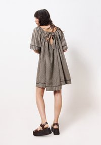 Schwarz-beige Gingham-Kleid mit kurzen Ärmeln, offener Rücken, der mit einem Band gesichert ist, und ausgestellter Silhouette. Kombiniert mit braunen Plateausandalen.
