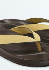 Tongs marron foncé avec larges brides en daim jaune moutarde et semelles moulées, photographiées sur un fond clair uni.