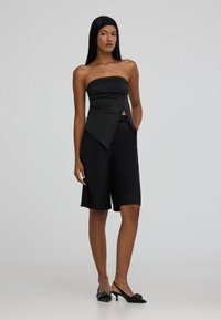 Top nero senza spalline con dettaglio anteriore attorcigliato, abbinato a pantaloni corti neri fino al ginocchio. Modella che indossa tacchi sling-back neri. Texture liscia.