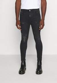 Hombre vistiendo jeans ajustados negros desgastados con manchas de pintura, botas de cuero negras y una camiseta blanca contra un fondo liso.