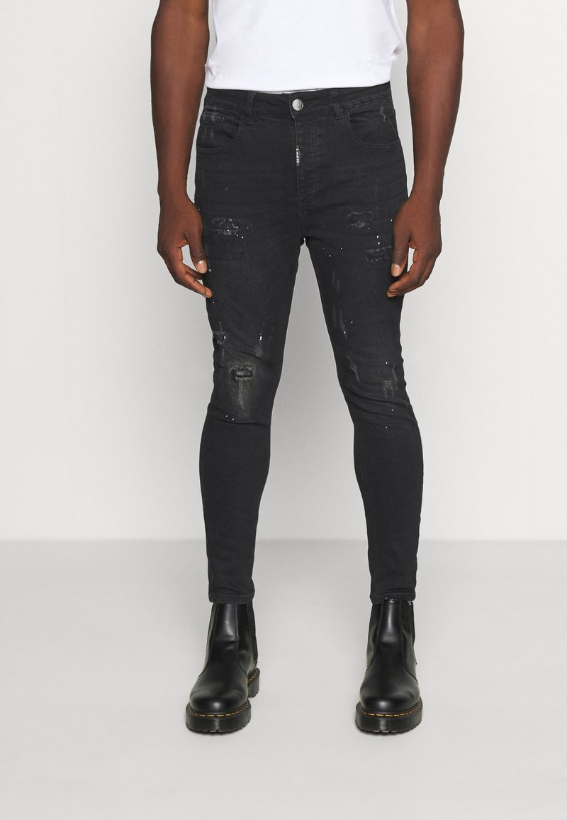 Hombre vistiendo jeans ajustados negros desgastados con manchas de pintura, botas de cuero negras y una camiseta blanca contra un fondo liso.