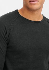 Chemise noire à manches longues en tissu tricoté texturé avec un col rond et des coutures raglan aux épaules.