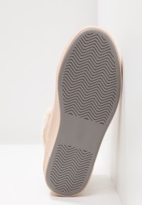 Semelle d'une chaussure beige avec une semelle extérieure en caoutchouc gris présentant un motif de traction en zigzag, reposant contre des surfaces blanches inclinées.